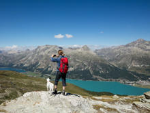 Eine Frau steht mit einem Hund auf einem Berg und macht mit ihrem Smartphone ein Bild von der Landschaft.